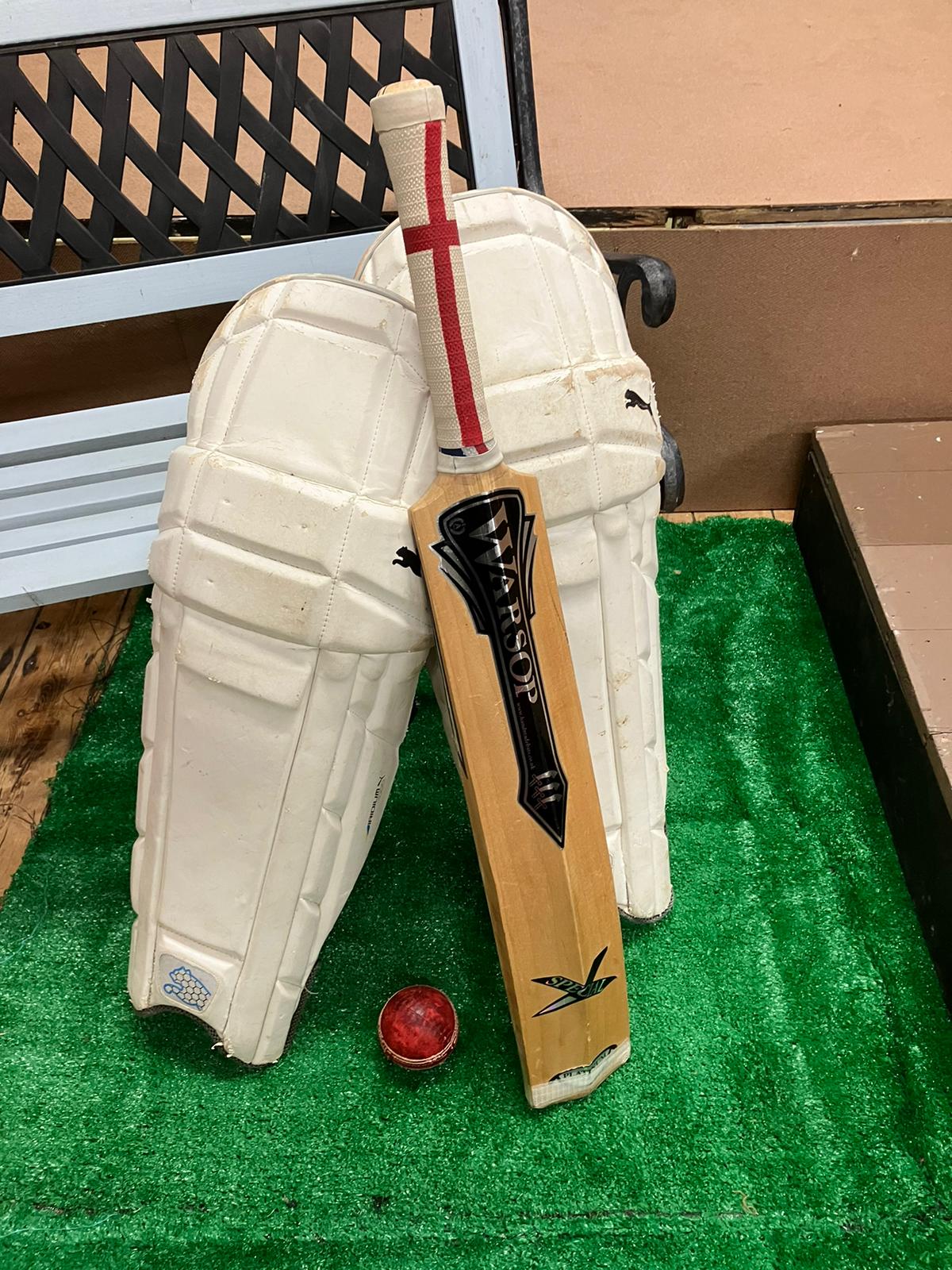A cluster of cricketing items - a ball, a bat and two cricket pads - is arranged on some artificial turf and leaning against a bench.